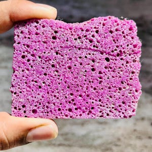 May include: A rectangular, bright magenta sponge with a porous, textured surface featuring numerous small, irregular holes distributed across the entire face of the material, showing a consistent density and a vibrant, saturated pink colour.