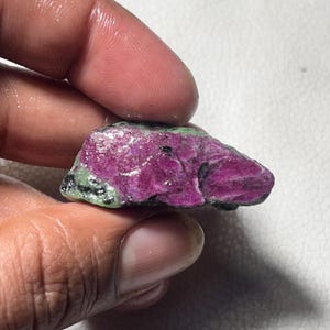 Könnte beinhalten: Ein roher Rubin-Edelstein mit tiefmagentafarbener Farbe, akzentuiert durch grüne und schwarze Flecken. Das rohe Mineral wird in einer Hand gehalten und zeigt seine natürliche, unpolierte Textur und lebendigen Farbvariationen.