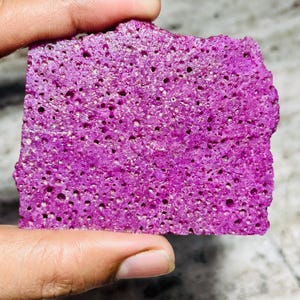 May include: A rectangular piece of porous, vibrant magenta material with a rough, uneven surface texture. The object is characterized by a dense network of small, circular holes distributed across its entire flat, front-facing surface area.
