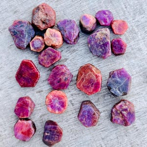 May include: A collection of raw ruby gemstones in various shapes and sizes. The stones display a range of colors, including deep reds, purples, and pinks, with some exhibiting a mix of hues. The rough texture and natural forms of the gems are visible.
