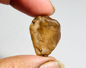 May include: A rough, amber-colored gemstone with a natural, irregular shape. The stone is held between two fingers against a white background, showcasing its translucent quality and subtle variations in color.