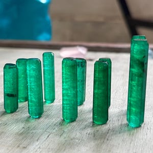 Op de afbeelding: Een verzameling levendige groene smaragdkristallen, elk met een gepolijste, cilindrische vorm. De edelstenen zijn gerangschikt op een lichtgekleurd oppervlak, waarbij hun rijke kleur en natuurlijke textuur worden benadrukt. De kristallen variëren in hoogte.