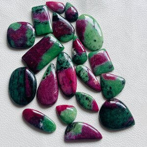 Può includere: Una collezione di pietre preziose di rubino zoisite lucide in varie forme, tra cui ovali, a goccia e rettangolari. Le pietre mostrano un mix di tonalità rosso intenso e verde, con una finitura lucida. Le pietre sono disposte su una superficie bianca.