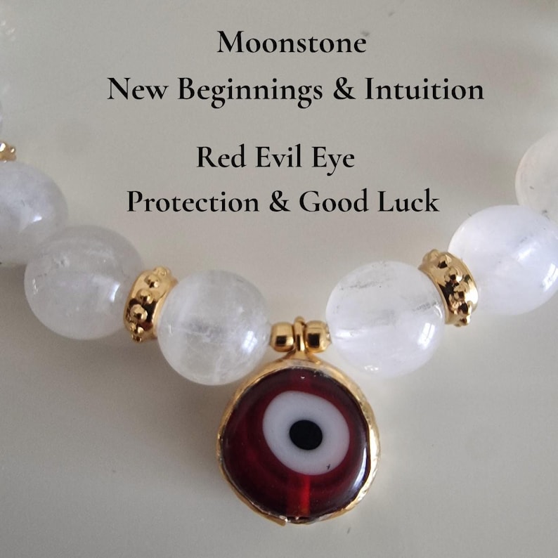May include: A beaded bracelet featuring translucent white moonstone beads, gold-toned metal spacers, and a central red evil eye charm. The text reads: Moonstone, New Beginnings & Intuition, Red Evil Eye, Protection & Good Luck.