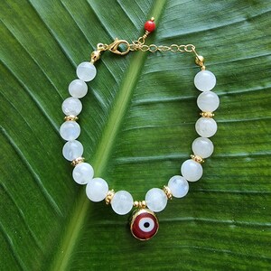 May include: A beaded bracelet featuring round white moonstone beads interspersed with small gold-toned metal spacers. The bracelet includes a central circular charm with a red, white, and black evil eye design and a small red bead at the clasp.