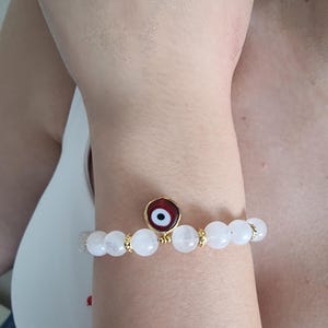 May include: A beaded bracelet featuring smooth, translucent white round beads and small gold-toned spacers. A circular charm with a red, white, and black evil eye design is positioned at the center of the bracelet, secured with gold-toned metal.