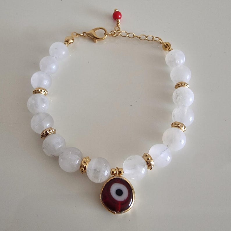 May include: A beaded bracelet featuring translucent white round beads interspersed with small gold-toned metal spacers. A circular charm with a red, white, and black eye design hangs from the center. The bracelet has a gold-toned chain clasp.