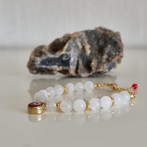 May include: A beaded bracelet featuring translucent white round beads, small gold-toned spacers, and a circular charm with a red, white, and blue evil eye design. The bracelet includes a gold-toned lobster clasp and a small red bead accent.