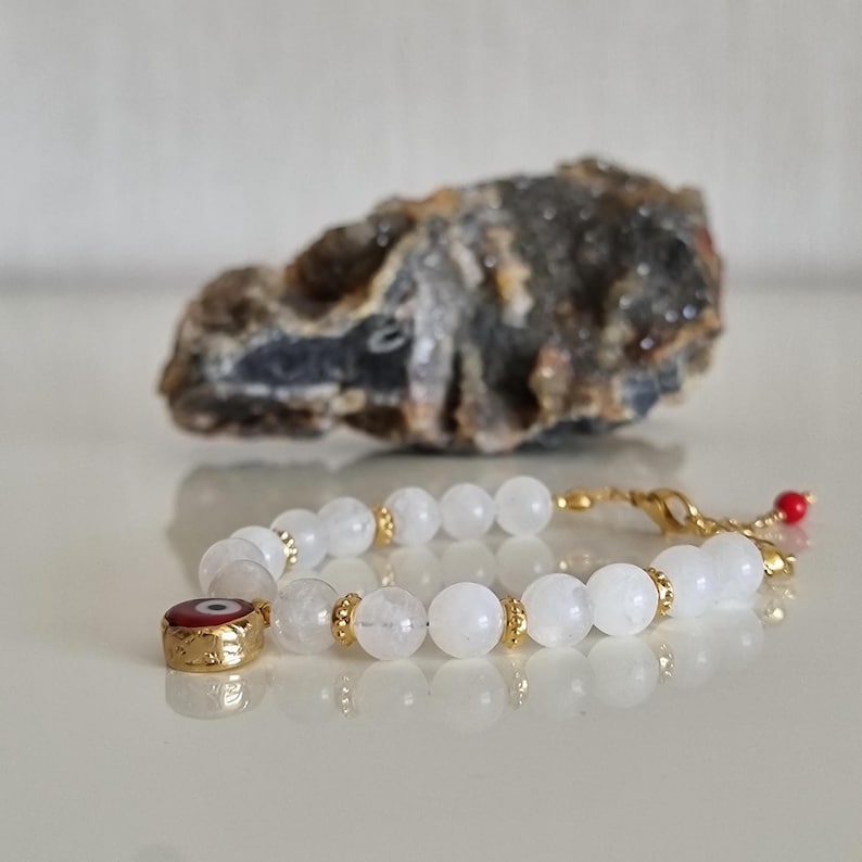 May include: A beaded bracelet featuring translucent white round beads, small gold-toned spacers, and a circular charm with a red, white, and blue evil eye design. The bracelet includes a gold-toned lobster clasp and a small red bead accent.