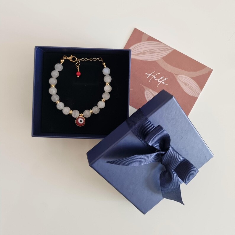 May include: A beaded bracelet with translucent white stones, gold-toned spacers, and a central red and white evil eye charm, presented in a dark blue gift box with a matching ribbon and a card featuring the word Hello in script.