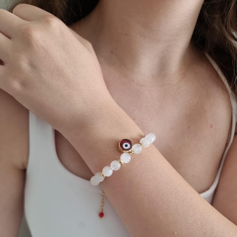 May include: A beaded bracelet featuring smooth, translucent white stones interspersed with small gold-toned metal spacers. A circular charm with a red, white, and black eye motif hangs from the center, and a gold chain with a red bead hangs below.