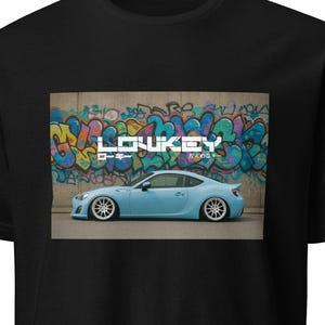 Peut inclure: T-shirt noir avec un graphisme d'une voiture de sport bleu clair devant un mur de graffitis. La voiture a des roues personnalisées et est abaissée. Le texte "LOWKEY" est au-dessus de la voiture.