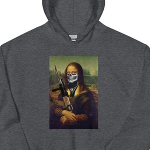 Puede incluir: Sudadera con capucha gris oscuro con una parodia de la Mona Lisa. La obra de arte representa a la Mona Lisa con una máscara de calavera y sosteniendo un rifle. La sudadera tiene una capucha con cordón y un bolsillo de canguro.
