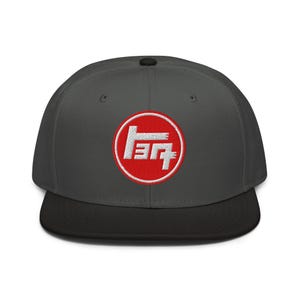 May include: A charcoal gray flat-brimmed baseball cap with a black brim. The cap features a red circular patch with white embroidered text. The text is a stylized logo.