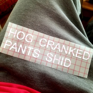 May include: A clear vinyl decal with the words "HOG CRANKED PANTS SHID" in white block letters. The decal is on a red grid backing, resting on gray fabric. A glimpse of red fabric is visible.