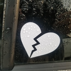 Può includere: Adesivo bianco a forma di cuore spezzato, con una crepa a zig-zag. L'adesivo è applicato su una superficie scura, probabilmente un finestrino dell'auto, ed è ricoperto di goccioline d'acqua. La forma del cuore è ben definita.