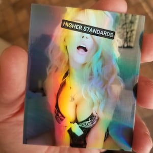 Può includere: Un adesivo olografico con il viso di una donna e il testo "HIGHER STANDARDS" sulla sua fronte. L'adesivo ha un effetto arcobaleno iridescente.
