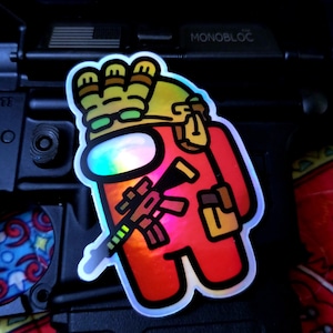 Op de afbeelding: Een holografische sticker met een rood stripfiguur met een kroon en zonnebril, die een geweer en een klein voorwerp vasthoudt. De sticker heeft een witte rand en is op een zwart oppervlak geplaatst.