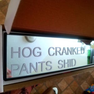 Puede incluir: Un letrero rectangular con las palabras "HOG CRANKED PANTS SHID" en gris, fuente sans-serif. El letrero está en un marco negro y dentro de una caja de cartón. El fondo es una superficie clara y reflectante.