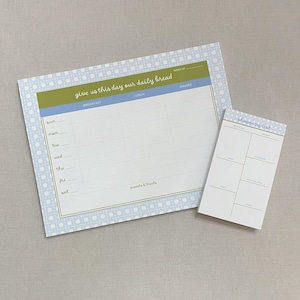 May include: A meal planning notepad and a shopping list notepad. The meal planner has a blue and green header with the text "give us this day our daily bread". The shopping list notepad has a blue header with the text "shopping list". Both have a light blue dotted border.