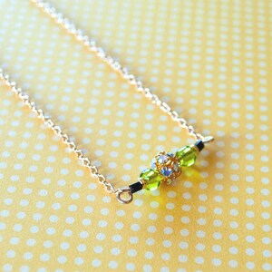 May include: A delicate gold-colored necklace with a central beaded design. The focal point features a cluster of iridescent and green beads, flanked by smaller green beads and black accents. The necklace rests on a yellow polka-dotted surface.