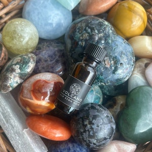 May include: A small brown glass bottle with a black label that reads "VOLCANO AIR SIGN OILS" is surrounded by a variety of colorful heart-shaped and round stones in a woven basket.