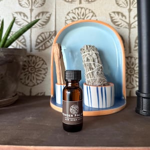 May include: A small brown glass bottle with a black cap and a label that reads "Tender Palo Air Sign Oil". The bottle is sitting on a wooden surface in front of a blue and white ceramic dish.