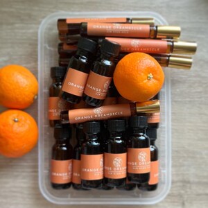 May include: An overhead shot shows a clear plastic container filled with small brown glass bottles and roll-ons. Each bottle has an orange label with the text "ORANGE DREAMSICLE". Two oranges sit outside the container. The roll-ons have gold-colored caps.