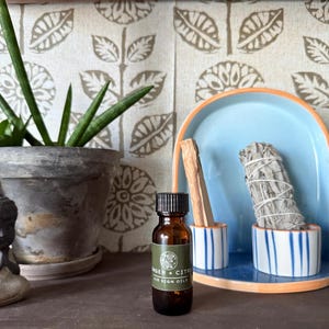 May include: A small brown glass bottle with a black cap and a white label that reads "Ginger + Citrus Air Sign Oil". The bottle is sitting on a wooden surface in front of a white background with a floral pattern. There is a sage smudge stick and a palo santo stick in the background.