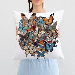 Puede incluir: Una almohada blanca con un diseño de mariposas de colores. Las mariposas están dispuestas en un patrón circular y son de varios tonos de azul, naranja, marrón y amarillo.