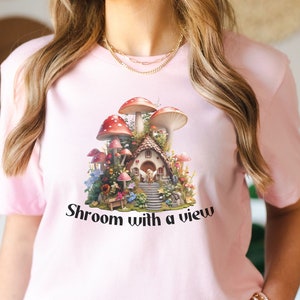 以下が含まれることがあります： きのこハウスと景色が描かれたピンクのTシャツ。グラフィックの下に「Shroom with a view」というテキストが印刷されています。