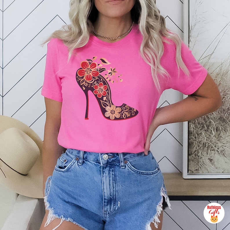 May include: A pink t-shirt featuring a floral design of a high-heeled shoe. The shoe is black with gold accents and red flowers.