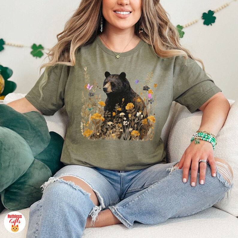 May include: A green t-shirt with a black bear sitting in a field of yellow flowers. The bear is surrounded by butterflies and other floral details.
