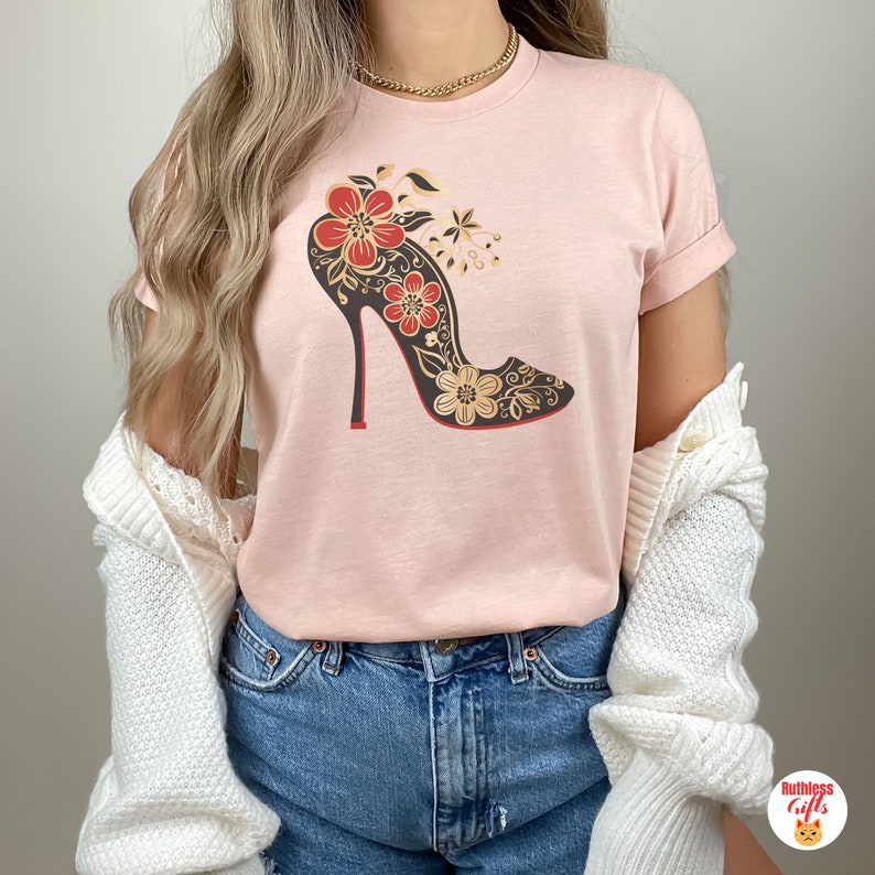 May include: A pink t-shirt featuring a black high-heeled shoe with red and gold floral details printed on the front.