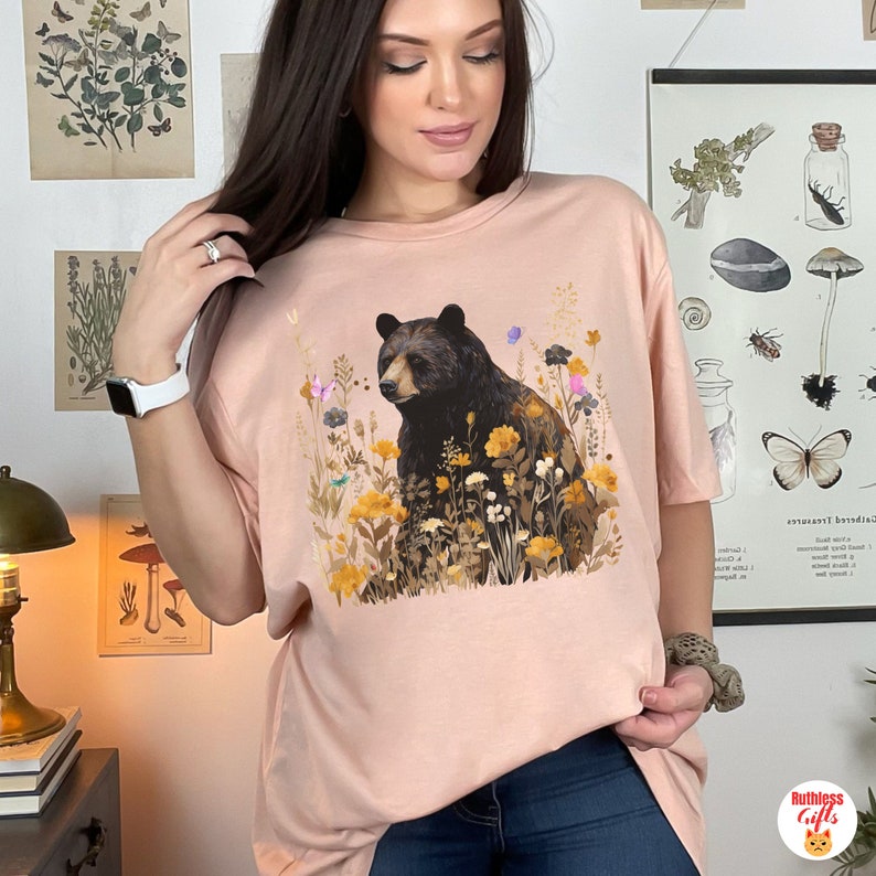 May include: A pink t-shirt with a black bear surrounded by yellow, orange, and purple flowers. The bear is looking to the left. The shirt has a relaxed fit.