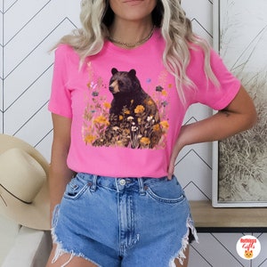 May include: A pink t-shirt with a black bear sitting in a field of flowers. The bear is surrounded by yellow, orange, and brown flowers. The shirt has short sleeves and a crew neck.