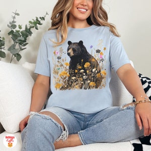 May include: A light blue t-shirt with a black bear illustration surrounded by yellow, orange, and pink flowers. The bear is looking to the left and has a brown nose.
