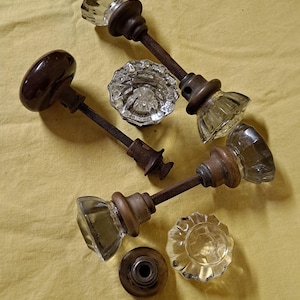 May include: An assortment of vintage door knobs and hardware. The knobs feature clear, faceted glass and dark brown wooden or metal bases. The hardware is arranged on a yellow fabric surface.
