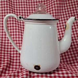 May include: White enamelware teapot with a clear glass lid and black trim. The teapot has a curved handle and spout. The background is a red and white checkered pattern. The teapot appears to be vintage or antique.