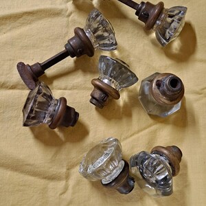 May include: A collection of vintage glass door knobs with faceted crystal-like designs. The knobs have aged bronze-colored metal bases and stems. The knobs are arranged on a yellow-tan fabric surface.