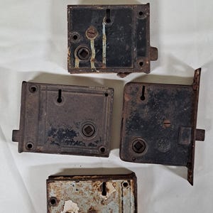 May include: Four rectangular metal mortise locks with visible signs of wear and oxidation. Each lock features a central square spindle hole, a keyhole, and mounting screw holes. The finishes include weathered black paint and rusted metal surfaces.