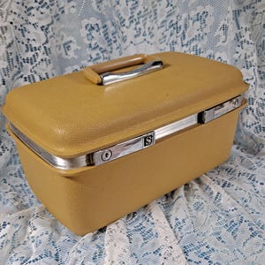 May include: Mustard yellow vintage suitcase with a textured surface, silver trim, and a handle. The suitcase has a rectangular shape and a metal clasp with the letter "S".