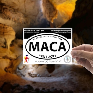 May include: White oval sticker with black text that reads "MAMMOTH CAVE NATIONAL PARK" and "MACA" in a larger font. Below the text is "KENTUCKY" and the coordinates "37.187004° N, 86.101183° W". The sticker is held in a hand against a blurry background of a cave.