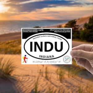 May include: White oval sticker with black text that reads "INDU Indiana Dunes National Park Indiana 41.638026° N, 87.095575° W". The sticker is being held by a hand against a blurred background of sand dunes and a sunset.