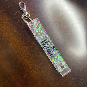 May include: A clear keychain with a silver clip and a pink and green glitter background. The keychain has the words "Be Brave" written on it.
