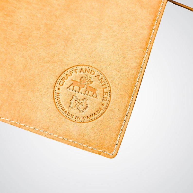 May include: A close-up of a light brown leather pouch with a stitched edge. The pouch has a circular logo embossed on the front that reads "Craft and Antler Handmade in Canada" with a stylized image of two deer and a C&A logo.