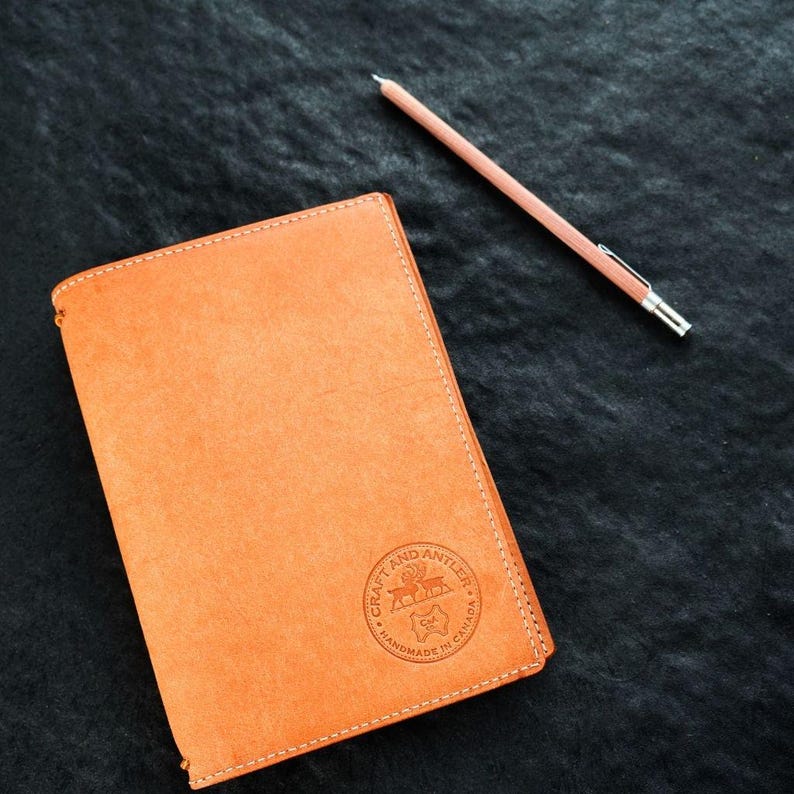 May include: A brown leather notebook with a stitched edge and a circular logo that reads "Craft and Antler Handmade in Canada". A pink pencil is lying on a black surface next to the notebook.