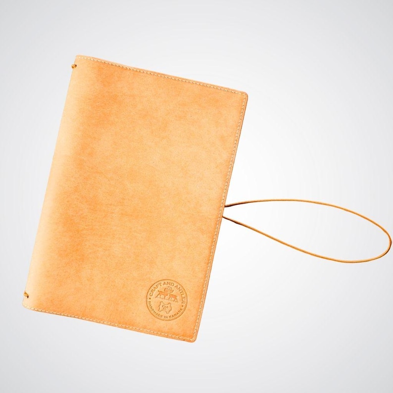 May include: A tan leather notebook cover with a brown leather strap and a circular logo that reads "Craft and Ancillary" with the text "Handcrafted in Canada" underneath.