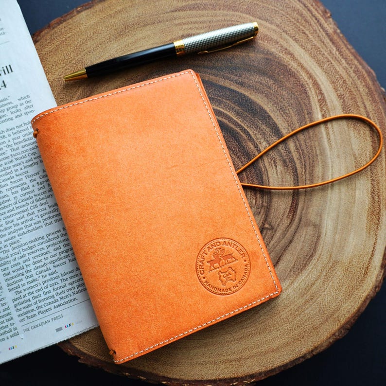 May include: A tan leather notebook cover with a stitched border and a brown leather strap. The cover has a circular embossed logo that reads "Craft and Antler Handmade in Canada".