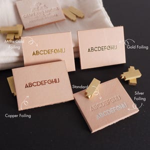 May include: Five leather labels with different foil finishes. The labels are all the same size and shape, and they all have the letters "ABCDEFGHIJ" printed on them. The foil finishes are: matte gold, gold, copper, silver, and standard.